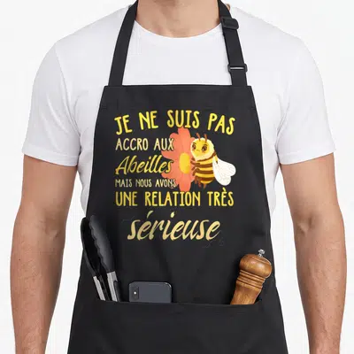 Tablier de cuisine noir avec une illustration d'abeille souriante tenant une fleur orange et un texte humoristique en jaune.