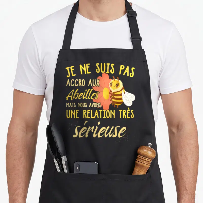 Tablier de cuisine noir avec une illustration d'abeille souriante tenant une fleur orange et un texte humoristique en jaune.