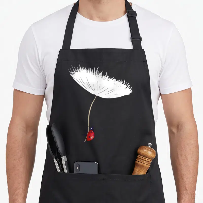 Tablier de cuisine noir avec une coccinelle rouge suspendue à une graine de pissenlit blanche stylisée.