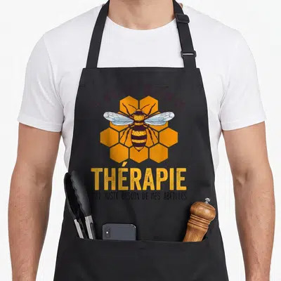 Tablier de cuisine noir avec une illustration d'abeille jaune et noire devant des motifs de ruche orange et du texte humoristique.