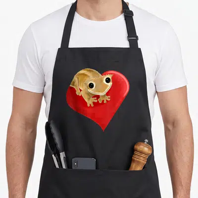 Tablier de cuisine noir avec illustration d'un lézard beige souriant devant un grand cœur rouge brillant.