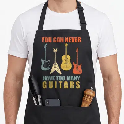 Tablier de cuisine noir avec quatre guitares vintage et l'inscription You Can Never Have Too Many Guitars imprimée en lettres colorées.