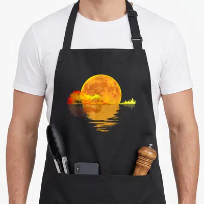 Tablier de cuisine noir avec illustration d'une lune orange, d'arbres automnaux colorés et leur reflet sur une surface aqueuse.