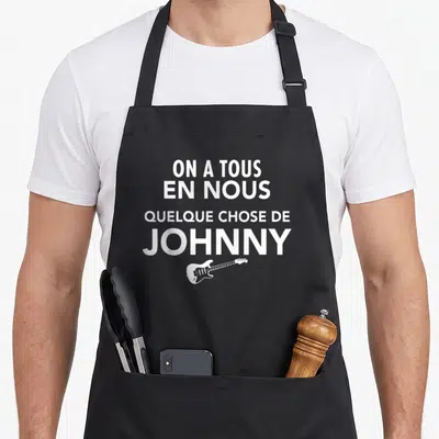 Tablier de cuisine noir avec le texte blanc On a tous en nous quelque chose de Johnny et une guitare électrique stylisée.