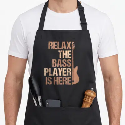 Tablier de cuisine noir avec texte Relax The Bass Player Is Here et silhouette de guitare basse orange.