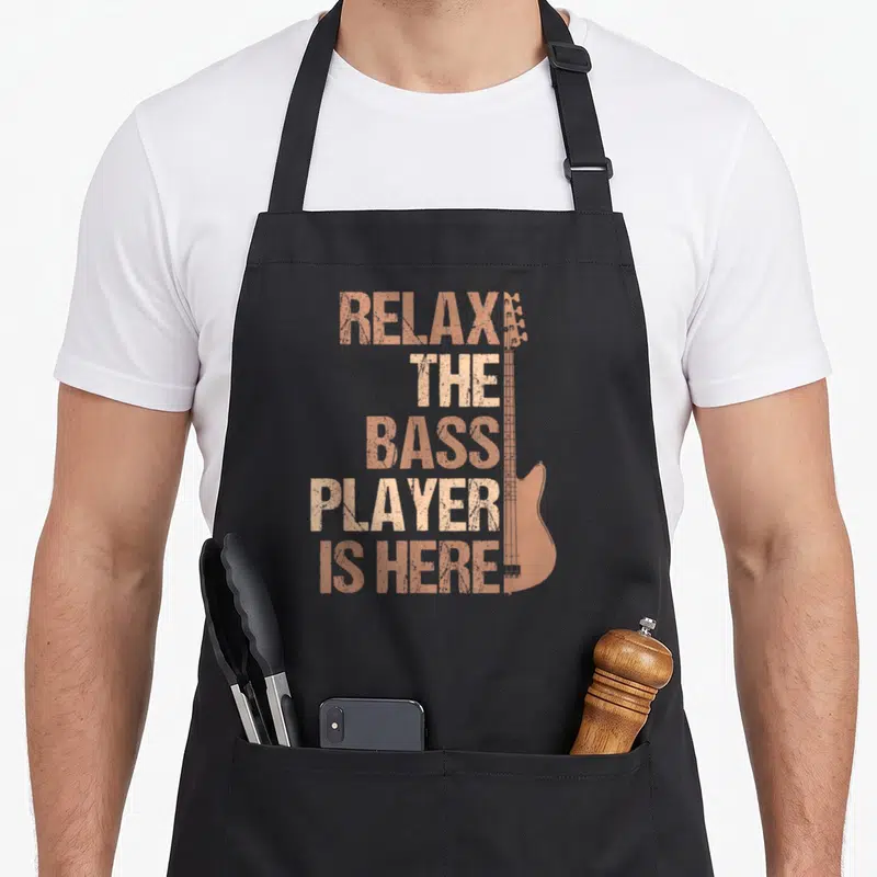 Tablier de cuisine noir avec texte Relax The Bass Player Is Here et silhouette de guitare basse orange.