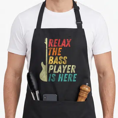 Tablier de cuisine noir avec silhouette de guitare basse et texte Relax The Bass Player Is Here en couleurs dégradées.