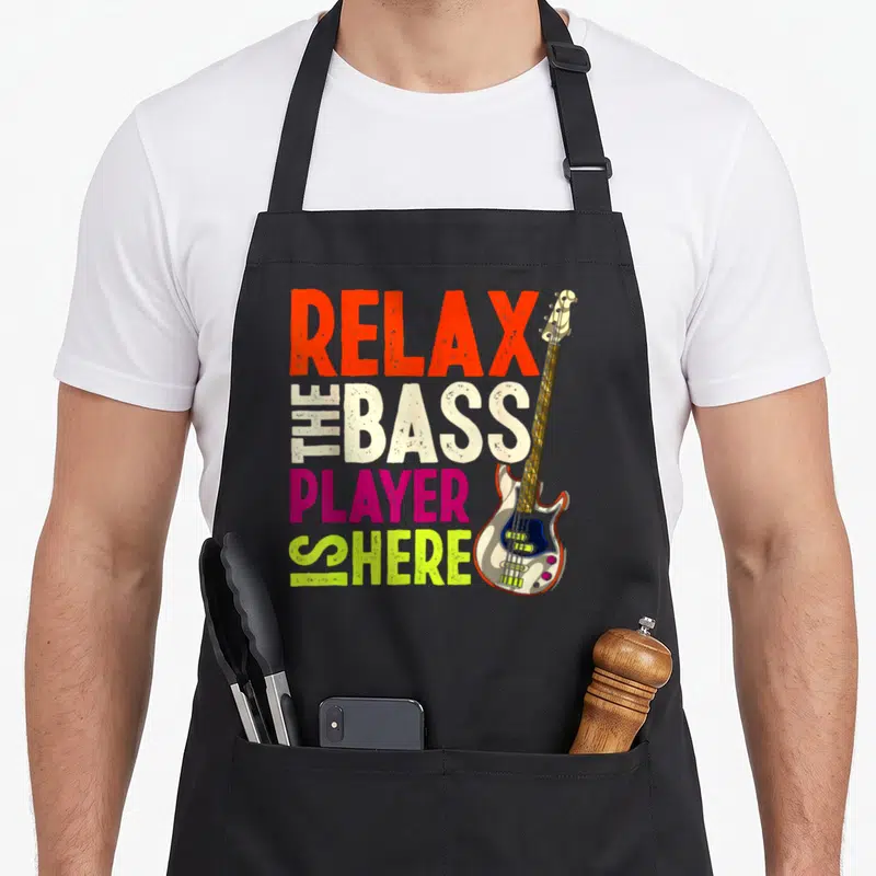 Tablier de cuisine noir avec texte multicolore Détendez-Vous Le Bassiste Est Ici et illustration de guitare basse.