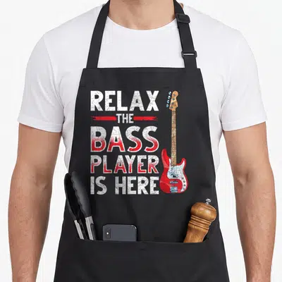 Tablier de cuisine noir avec texte Détendez-Vous Le Bassiste Est Ici et une guitare basse rouge à côté.