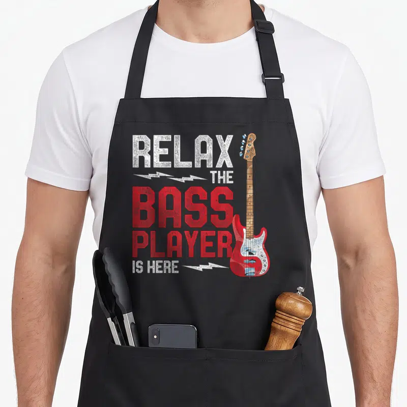 Tablier de cuisine noir avec inscription Détendez-Vous Le Bassiste Est Ici et illustration de guitare basse rouge à côté du texte.