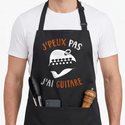 Tablier de cuisine noir avec illustration tête de mort barbu et texte J'peux pas j'ai guitare en orange et blanc.
