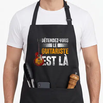 Tablier de cuisine noir avec illustration guitare électrique rouge et texte détendez-vous le guitariste est là en blanc et orange.