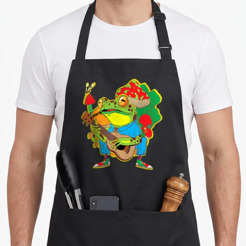 Tablier de cuisine noir avec une grenouille verte jouant de la guitare sur des champignons rouges dans un style graphique coloré.