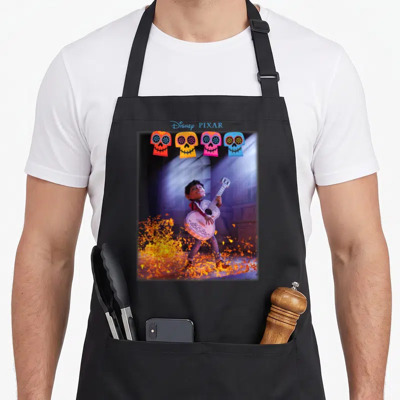 Tablier de cuisine noir avec Coco Miguel jouant d'une guitare blanche sous des projecteurs et quatre masques de calaveras colorés en haut.