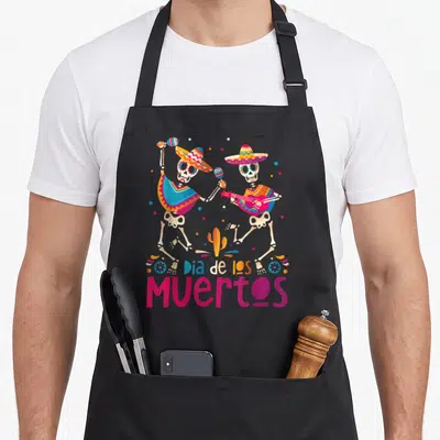 Tablier de cuisine noir avec deux squelettes mexicains dansant et jouant d'une guitare rose sous le texte Dia de los Muertos.