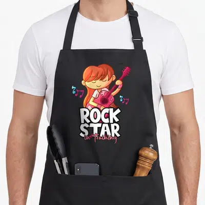 Tablier de cuisine noir avec illustration d'une fille guitariste aux cheveux roux et texte Rock Star in Training en blanc et rose.