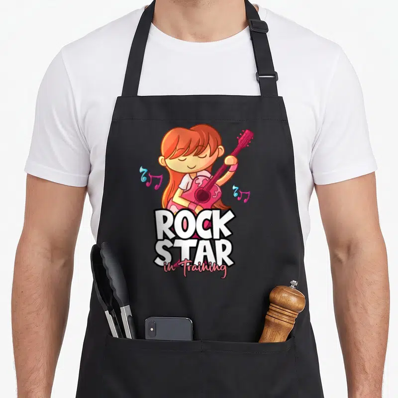 Tablier de cuisine noir avec illustration d'une fille guitariste aux cheveux roux et texte Rock Star in Training en blanc et rose.