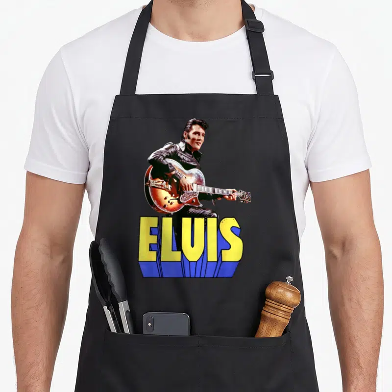 Tablier de cuisine noir avec portrait d'Elvis Presley jouant de la guitare et son nom en grosses lettres colorées.