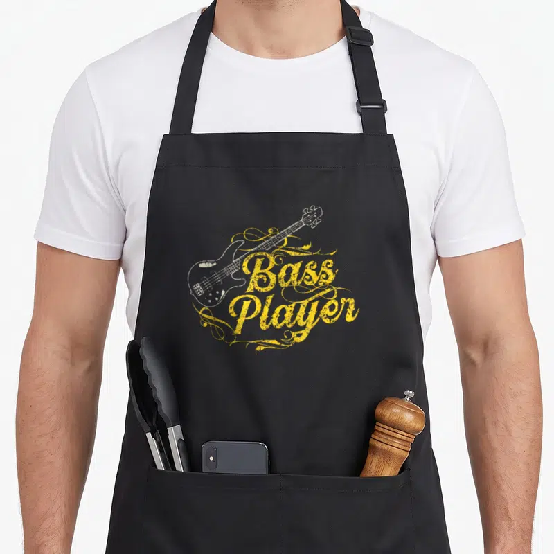 Tablier de cuisine noir avec illustration d'une basse électrique et le texte « Bass Player » en typographie jaune ornée de volutes.