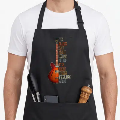 Tablier de cuisine noir avec une guitare blues orange et un texte humoristique en lettres colorées sur le côté droit.