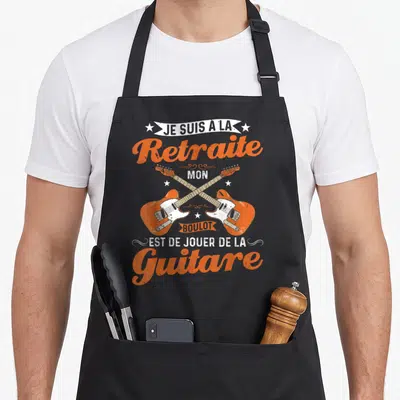 Tablier de cuisine noir avec deux guitares électriques oranges croisées et le texte Je suis à la retraite mon boulot est de jouer de la guitare.