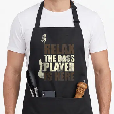 Tablier de cuisine noir avec texte Relax the bass player is here et silhouettes de guitares basse en style vintage.