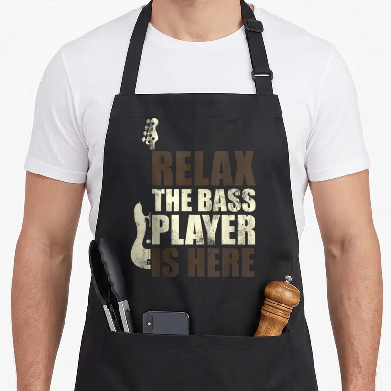 Tablier de cuisine noir avec texte Relax the bass player is here et silhouettes de guitares basse en style vintage.
