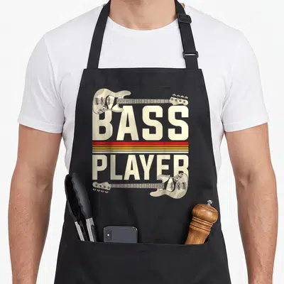 Tablier de cuisine noir avec illustration vintage BASS PLAYER et deux guitares basses beige encadrant des bandes rouges et jaunes.