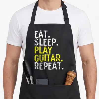 Tablier de cuisine noir avec inscription blanche et jaune EAT SLEEP PLAY GUITAR REPEAT en typographie délavée.