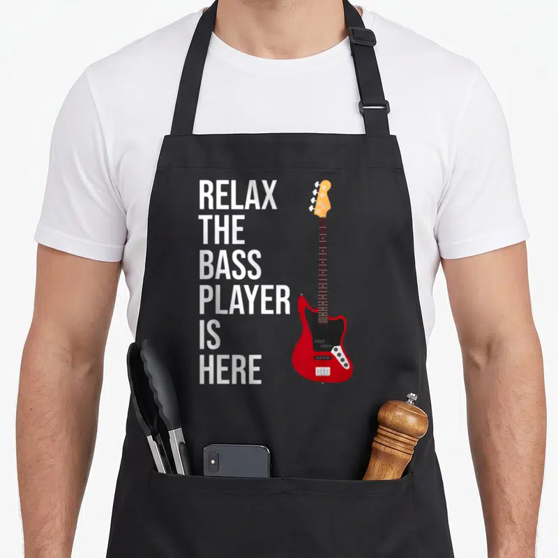 Tablier de cuisine noir avec une illustration de basse rouge et le texte Relax The Bass Player Is Here en blanc.