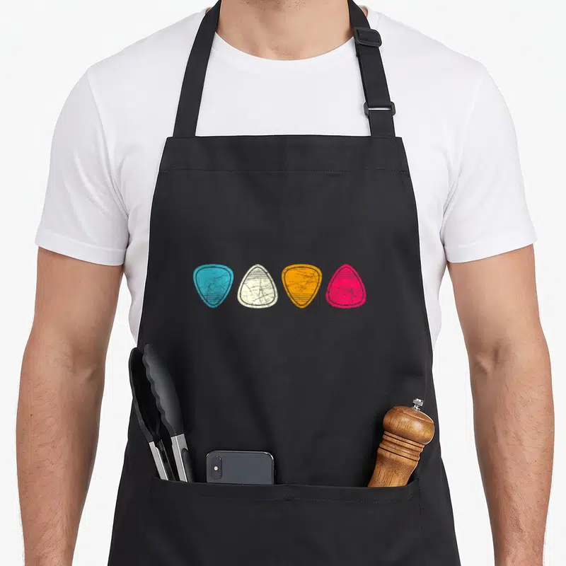 Tablier de cuisine noir avec quatre médiators de guitare colorés et usés alignés au centre.