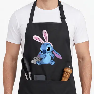 Tablier de cuisine noir avec illustration de Stitch portant un masque de lapin rose et tenant un petit animal gris.