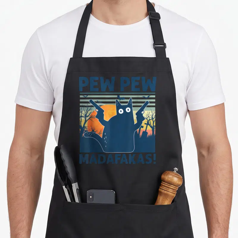 Tablier de cuisine noir avec illustration d'un chat tenant deux pistolets devant un soleil orange et des arbres morts, texte PEW PEW MADAFAKAS en haut et bas.
