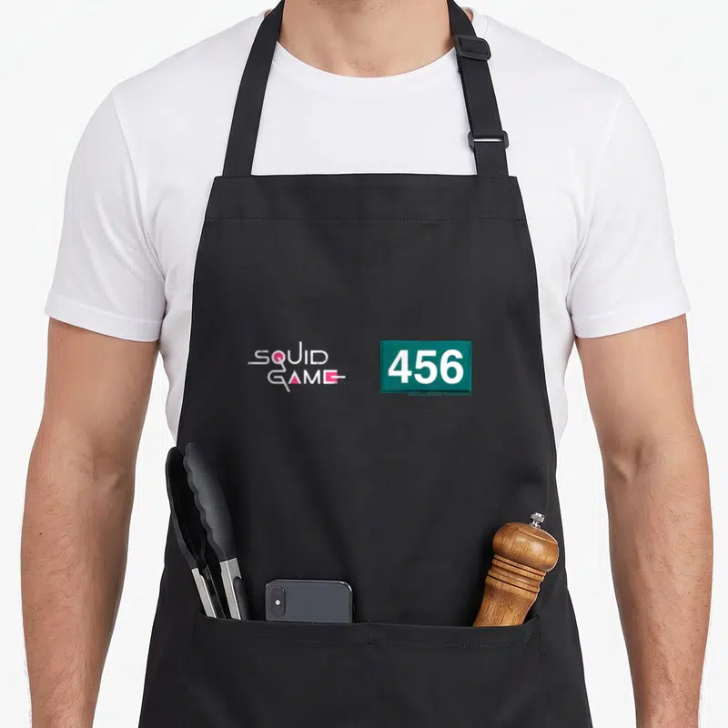 Tablier de cuisine noir avec logo Squid Game et numéro 456 sur fond vert émeraude.