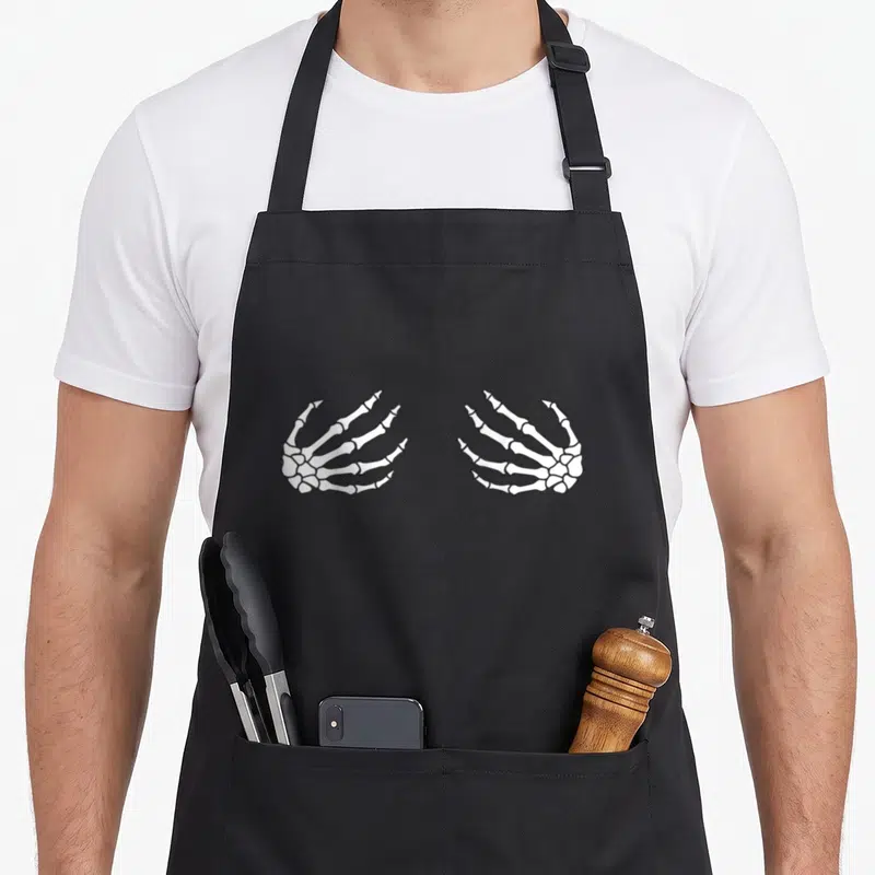 Tablier de cuisine noir avec deux mains squelette blanches dessinées au centre.