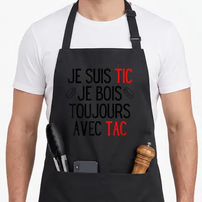 Tablier de cuisine noir avec l'inscription Je suis Tic Je bois toujours avec Tac en noir et rouge, flanquée de deux bouchons de bière.