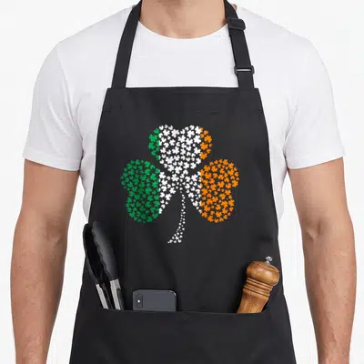Tablier de cuisine noir avec un grand trèfle formé de multiples petits shamrocks verts blancs et oranges sur le devant.