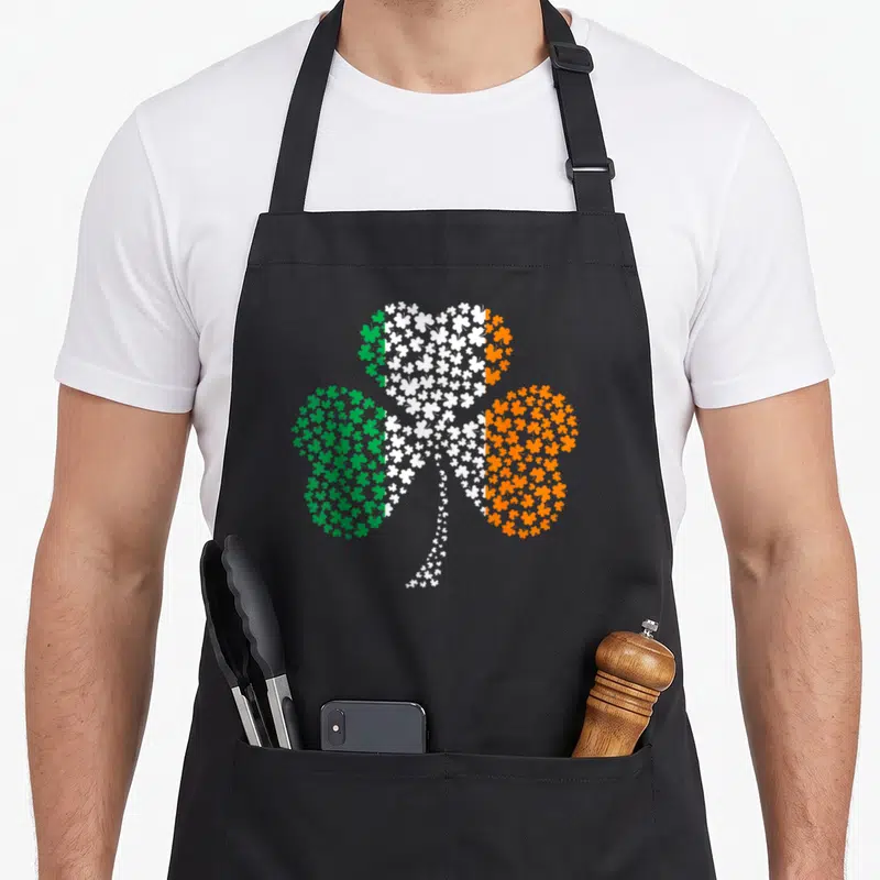 Tablier de cuisine noir avec un grand trèfle formé de multiples petits shamrocks verts blancs et oranges sur le devant.