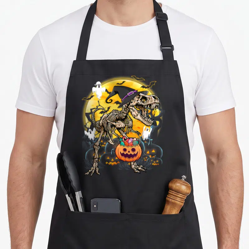 Tablier de cuisine noir avec un squelette de dinosaure T Rex portant un chapeau de sorcière et tenant une citrouille remplie de bonbons.