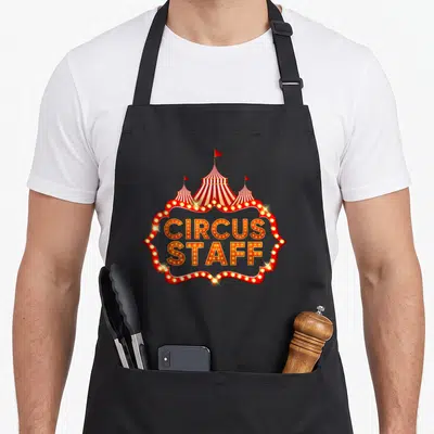 Tablier de cuisine noir avec une enseigne lumineuse Circus Staff et une tente de cirque rouge et blanche.