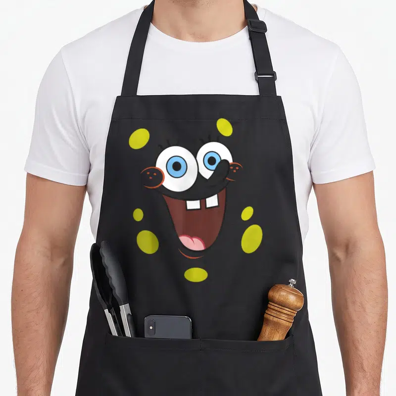 Tablier de cuisine noir avec un grand visage souriant de Spongebob aux yeux bleus et taches jaunes sur fond gris.