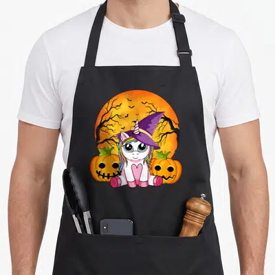 Tablier de cuisine noir avec une licorne sorcière assise entre deux pumpkins sur un fond de lune orange.