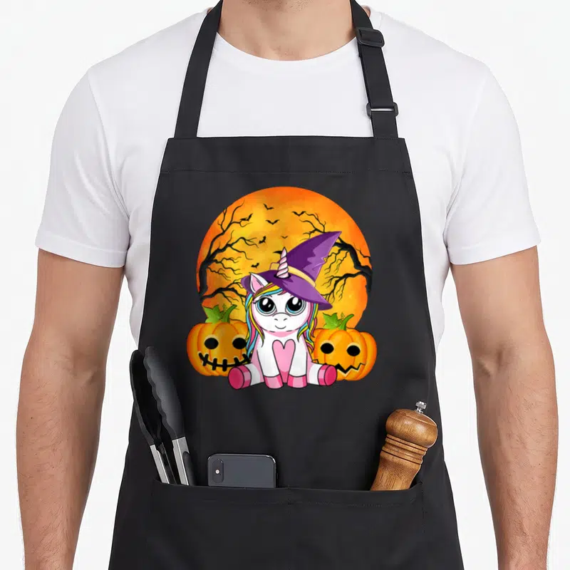 Tablier de cuisine noir avec une licorne sorcière assise entre deux pumpkins sur un fond de lune orange.