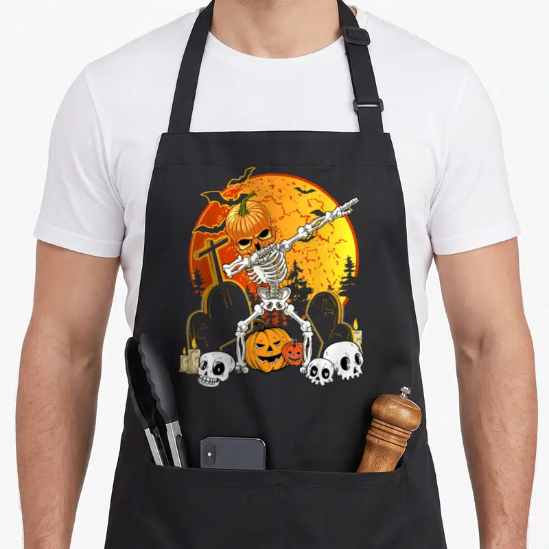 Tablier de cuisine noir avec squelette en train de faire le dab au milieu d'un cimetière et de citrouilles.