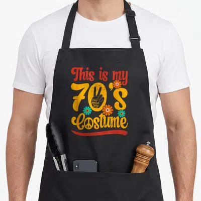 Tablier de cuisine noir avec texte This is my 70's Costume, main peace, fleurs colorées et Love peace joy en typographie vintage.