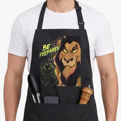 Tablier de cuisine noir avec illustration colorée du lion Scar et texte Be Prepared jaune vif.