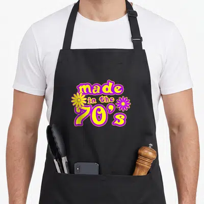 Tablier de cuisine noir avec inscription « made in the 70s » en jaune et violet entouré de deux fleurs stylisées.