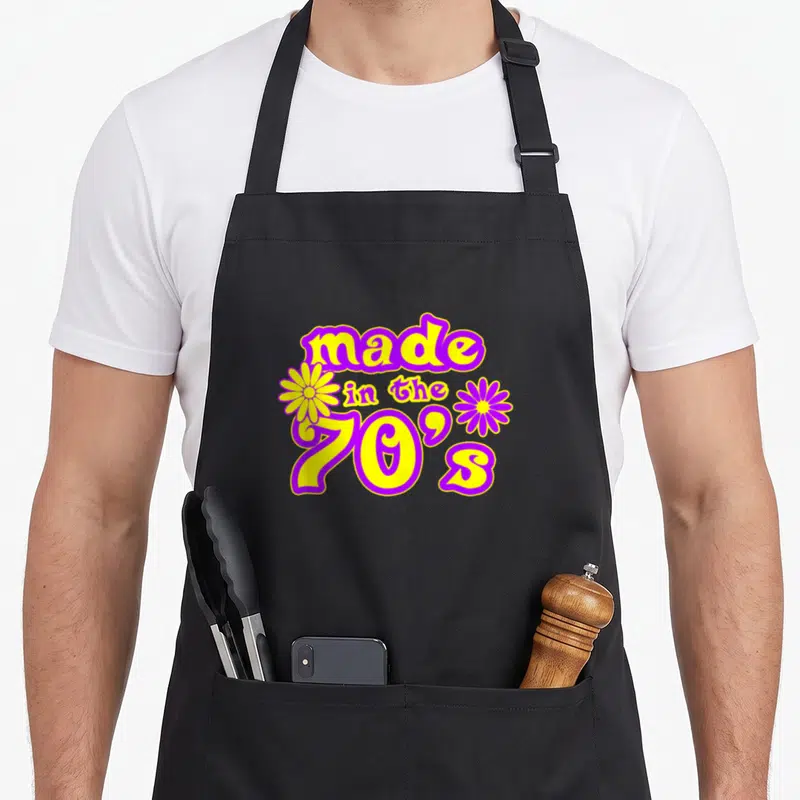 Tablier de cuisine noir avec inscription « made in the 70s » en jaune et violet entouré de deux fleurs stylisées.