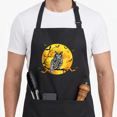 Tablier de cuisine noir avec une chouette blanche sur une lune orange entourée de chauves-souris noires.