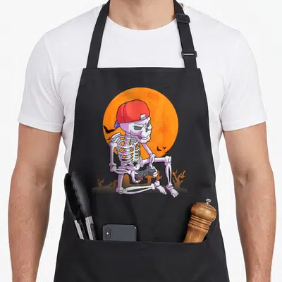 Tablier de cuisine noir avec squelette gamer portant une casquette rouge et tenant une manette de jeu devant un disque solaire orange.